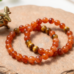 Carnelian and tiger's eye crystal bead bracelets on terracotta slate beside a brass ram figurine
