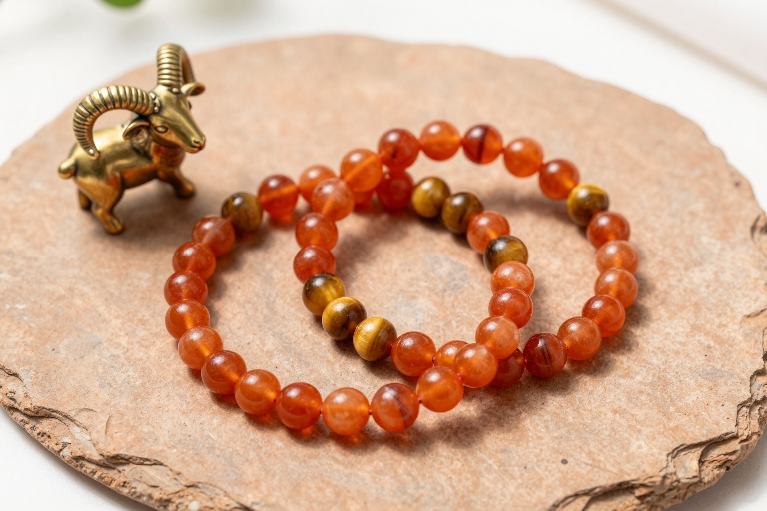 Carnelian and tiger's eye crystal bead bracelets on terracotta slate beside a brass ram figurine