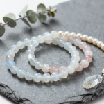 Moonstone, blue chalcedony and rose quartz crystal bead bracelets layered with a pearl strand and a raw selenite crystal on a grey slate slab beside dried eucalyptus, flat-lay in soft natural light