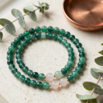 Emerald, rose quartz and jade bead bracelets arranged on cream travertine beside a copper dish and eucalyptus sprigs