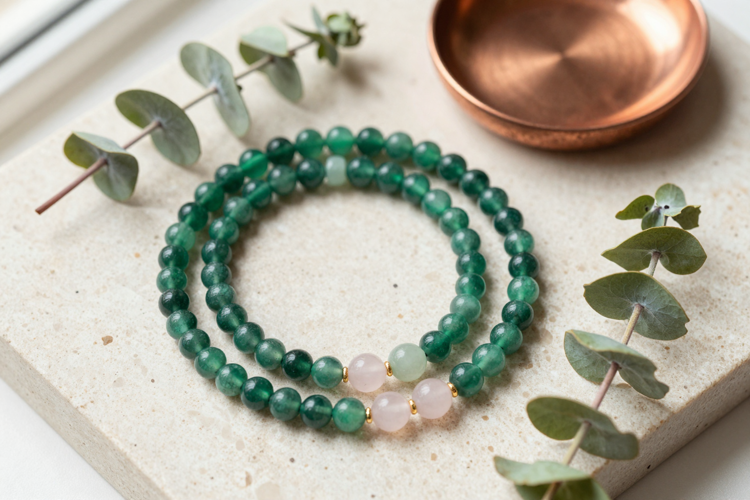 Emerald, rose quartz and jade bead bracelets arranged on cream travertine beside a copper dish and eucalyptus sprigs
