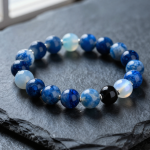 Handmade water element crystal bead bracelet with deep blue sodalite, blue lace agate, glossy black obsidian and moonstone beads, flat-lay on dark slate with water droplets