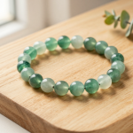 Handmade wood element crystal bead bracelet with green aventurine, jade and moss agate beads, flat-lay on light oak wood with a eucalyptus sprig, soft natural light