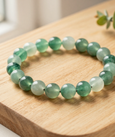 Handmade wood element crystal bead bracelet with green aventurine, jade and moss agate beads, flat-lay on light oak wood with a eucalyptus sprig, soft natural light